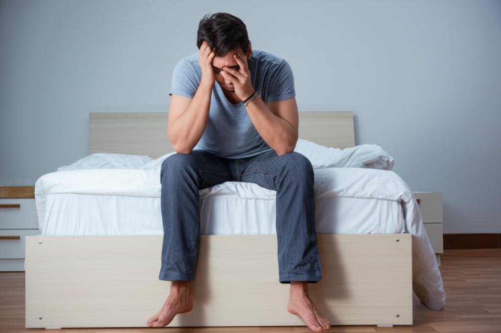 A sad looking man sitting on the edge of the bed, feeling frustrated and disappointed.