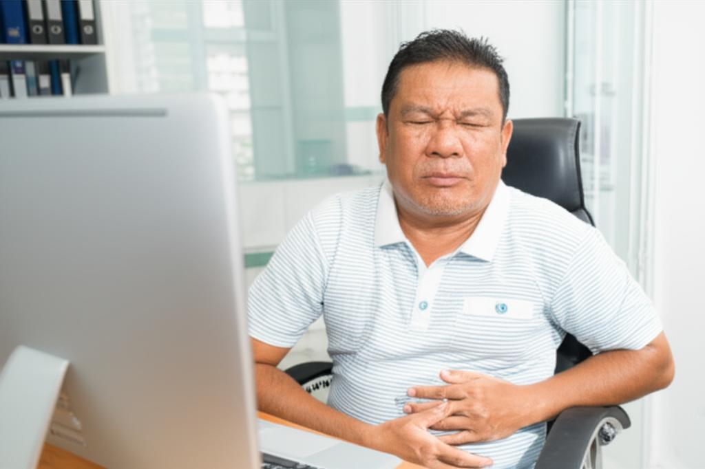 Middle-aged Malaysian man sitting at a desk, looking uncomfortable and distracted, holding his lower abdomen, office environment